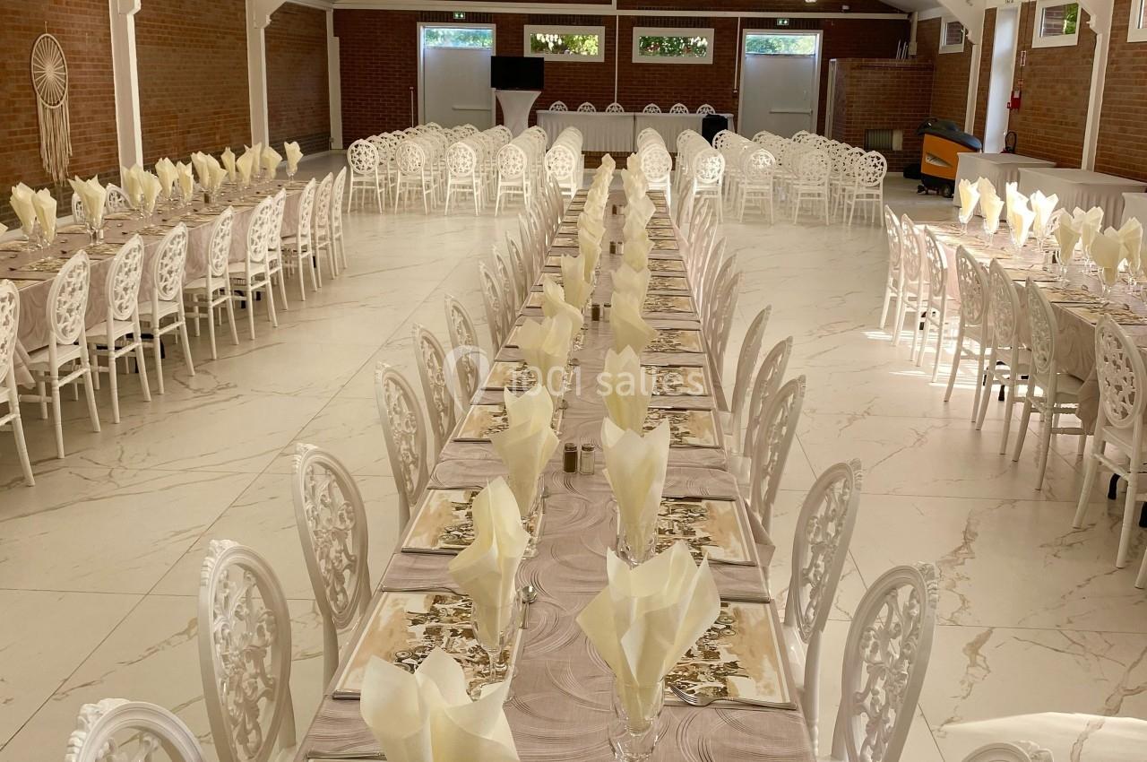 Salle de réception avec de longues tables dressées, nappes beige, serviettes pliées et chaises blanches décoratives.