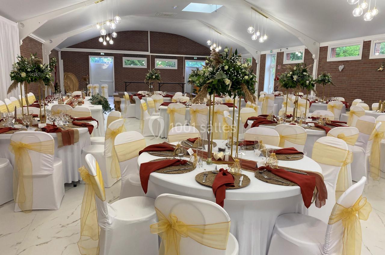 Salle de réception décorée pour un événement, avec des tables rondes ornées de nappes blanches, centres de table fleuris et…