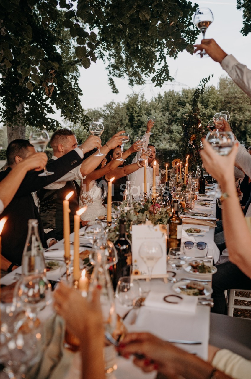 Des convives trinquent autour d'une longue table décorée de bougies et de fleurs lors d'un repas en plein air.