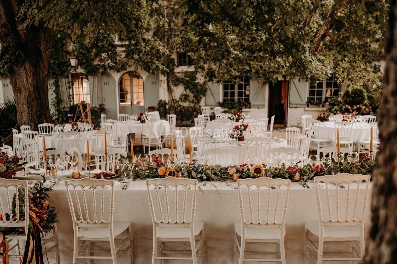 Tables de réception décorées avec des fleurs et des bougies, disposées en extérieur sous des arbres feuillus.