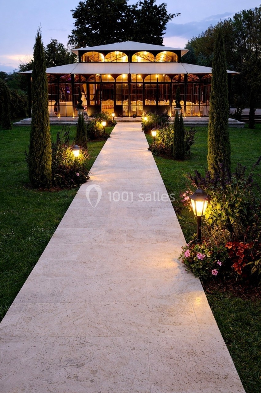 Allée pavée éclairée menant à un bâtiment vitré entouré de verdure au crépuscule.