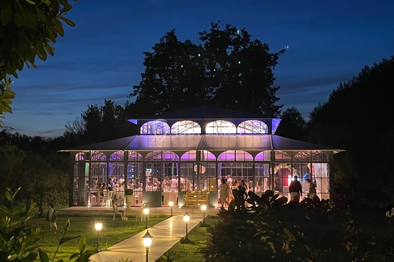 Serre illuminée en soirée, entourée de végétation et éclairée par des lampes le long d'un chemin.