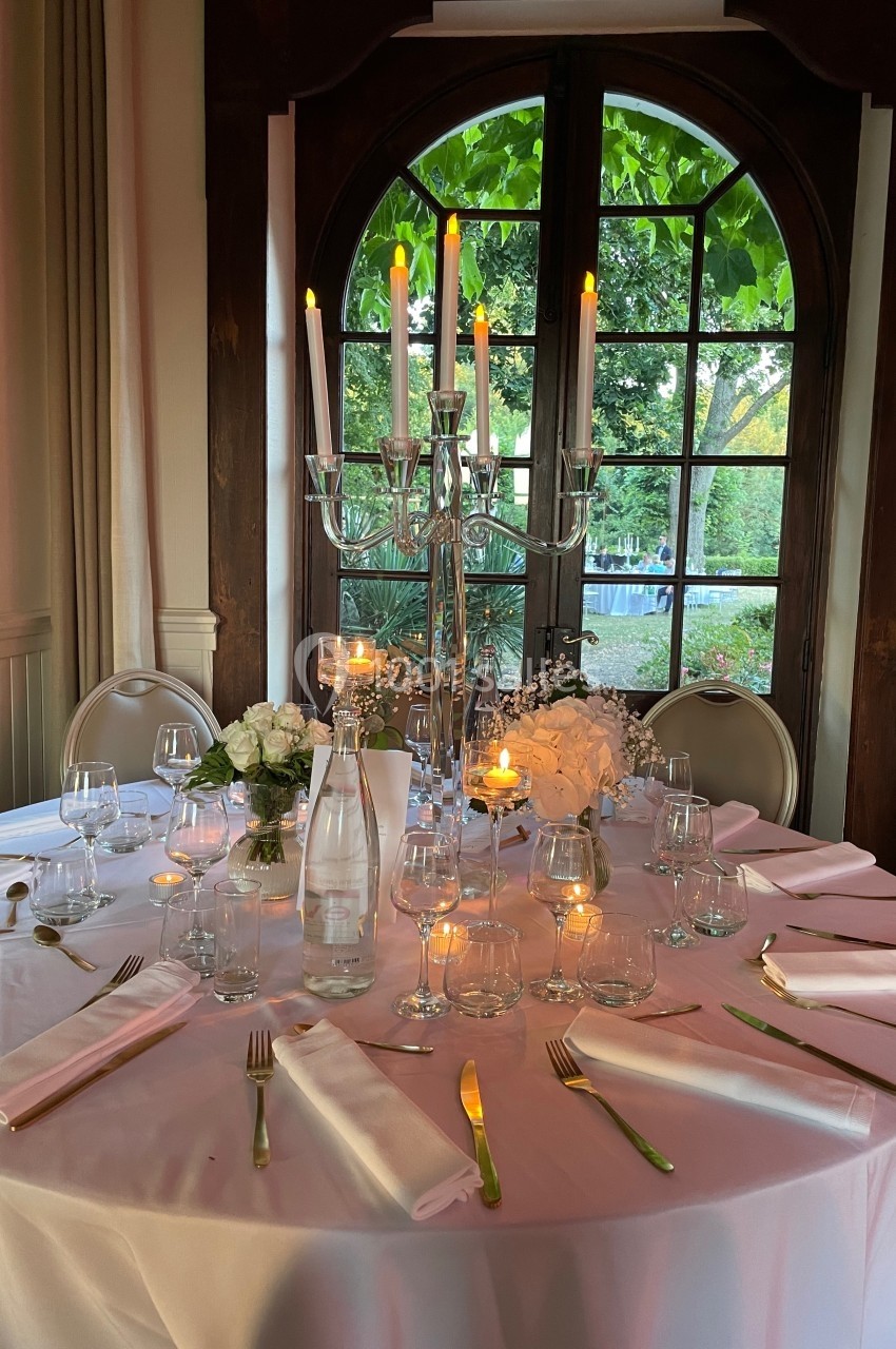 Table élégamment dressée avec chandelier, vaisselle raffinée et fleurs blanches, devant une fenêtre donnant sur un jardin.