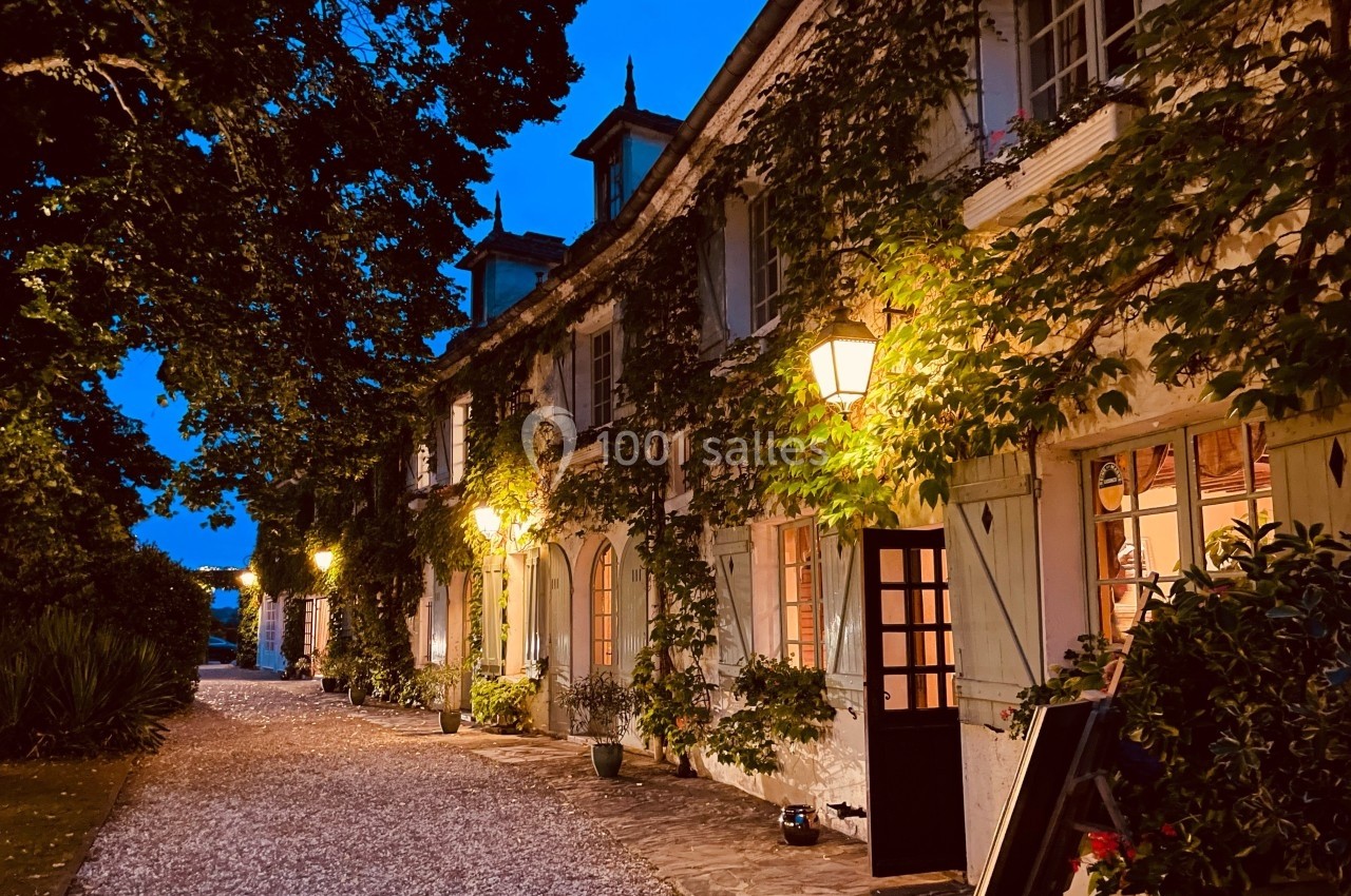 Façade d'une maison ancienne couverte de lierre, éclairée par des lampadaires au crépuscule, avec une allée en gravier.