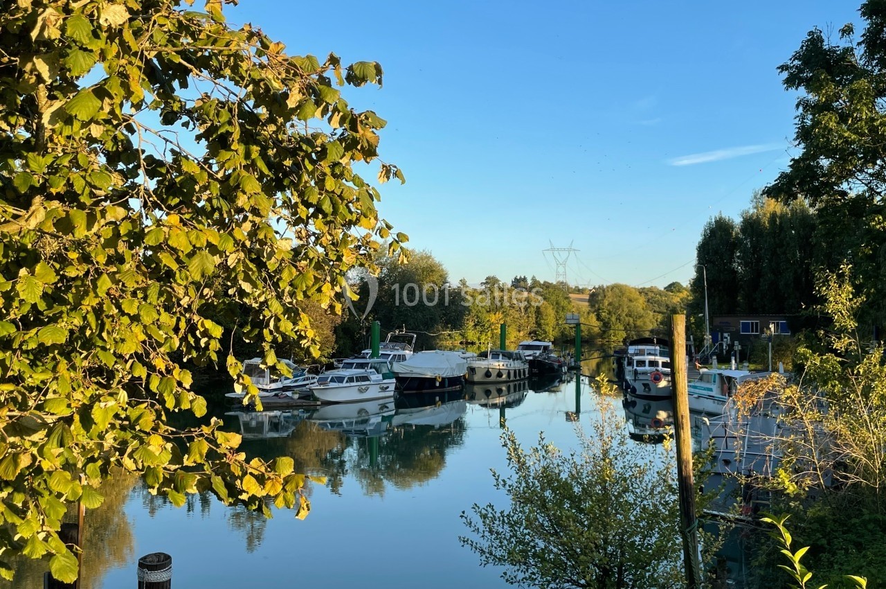 Canal bordé d'arbres avec des bateaux amarrés, reflétés dans l'eau calme sous un ciel bleu clair.