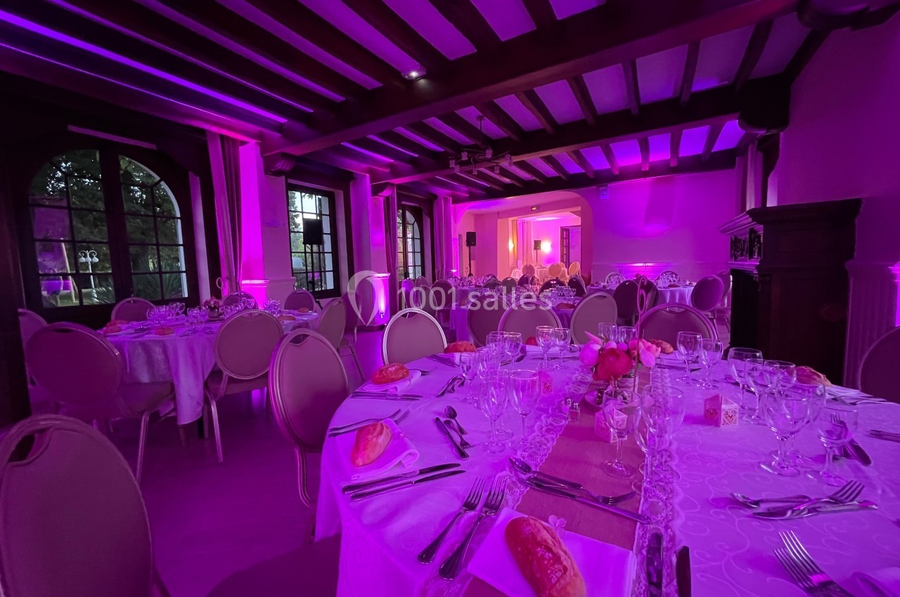 Salle de réception élégamment décorée avec des tables rondes dressées, éclairage rose et poutres apparentes.