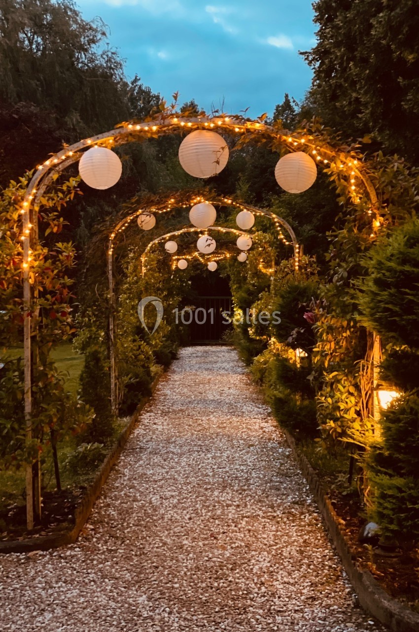 Allée de gravier bordée d'arches décorées de guirlandes lumineuses et de lanternes en papier, au crépuscule.