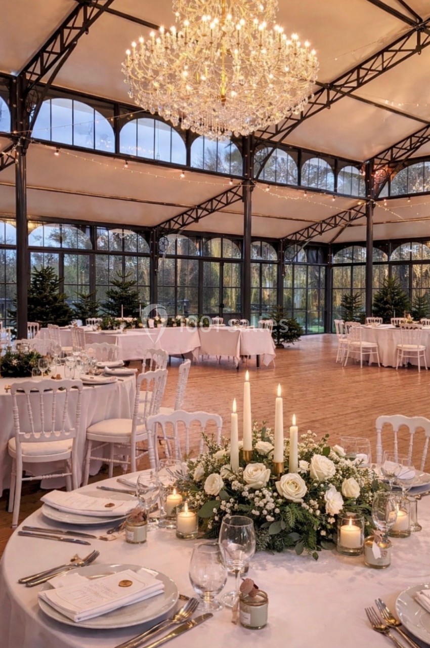 Salle de réception élégante avec tables décorées de fleurs blanches, bougies et grand lustre suspendu.