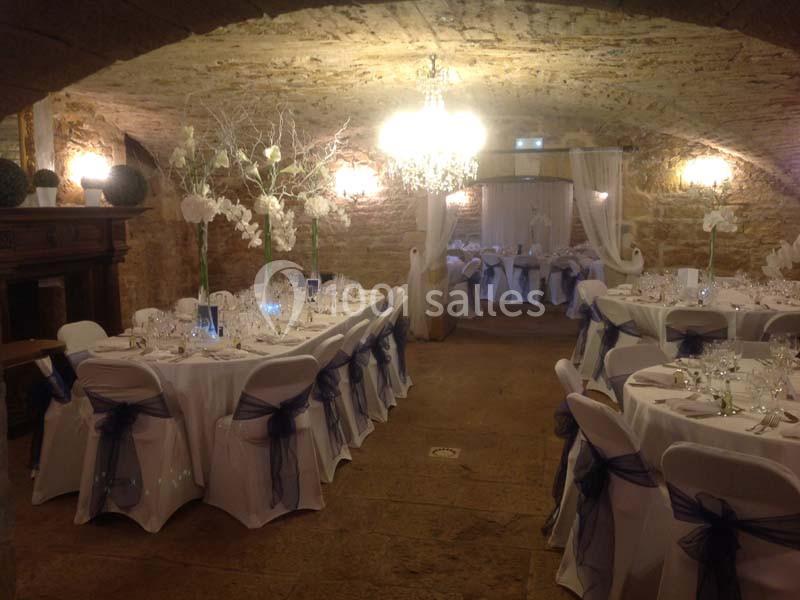 Salle voûtée décorée pour un mariage, avec tables rondes dressées, chaises blanches et éclairage chaleureux.