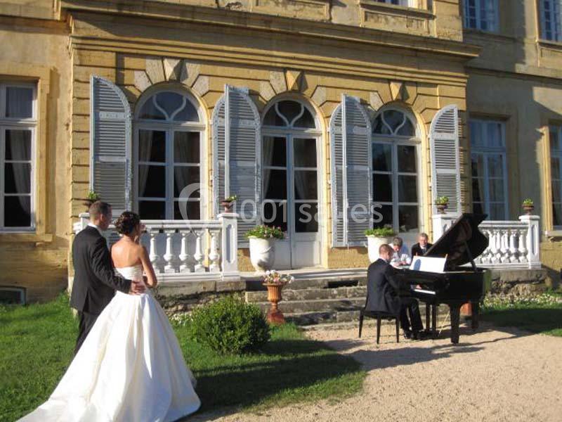 Un couple de mariés regarde un pianiste jouer devant une grande maison avec des fenêtres à volets ouverts.