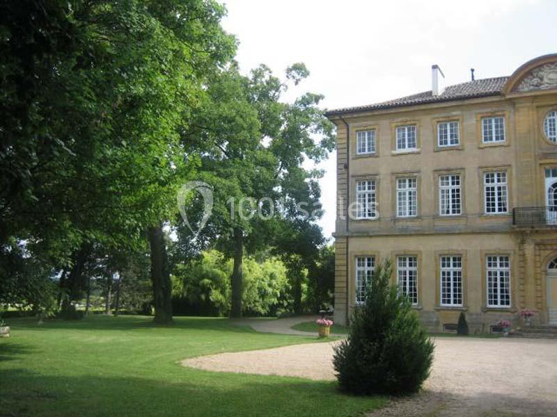 Façade d'un bâtiment ancien en pierre entouré d'un parc arboré avec pelouse et allée gravillonnée.