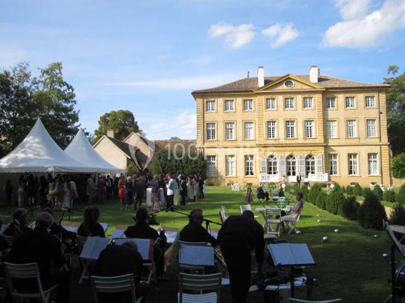 Jardin d'un château avec des invités rassemblés sous des tentes blanches et un orchestre en plein air au premier plan.