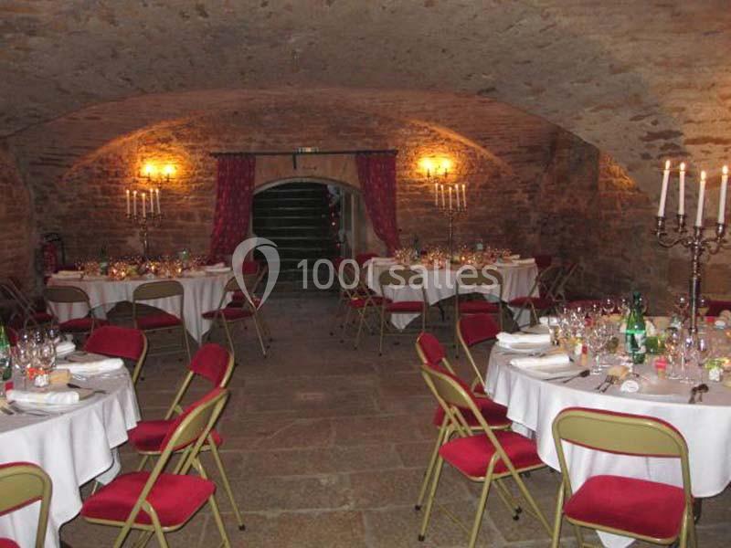 Salle voûtée en pierre aménagée pour un repas, avec tables rondes dressées, chaises rouges et chandeliers.