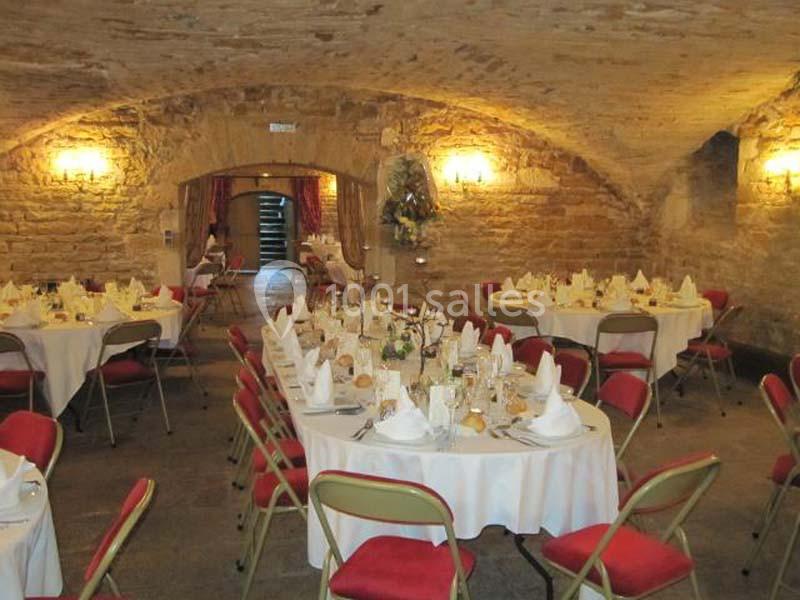 Salle voûtée en pierre aménagée pour un repas, avec tables rondes dressées et chaises rouges.