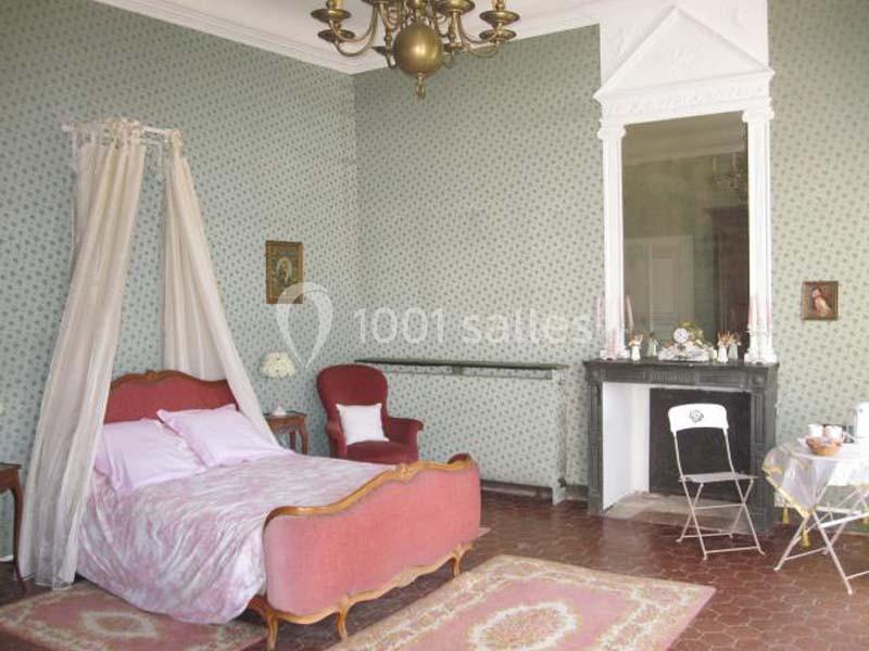 Chambre ancienne avec lit à baldaquin, fauteuil rouge, cheminée noire et murs tapissés de motifs verts.