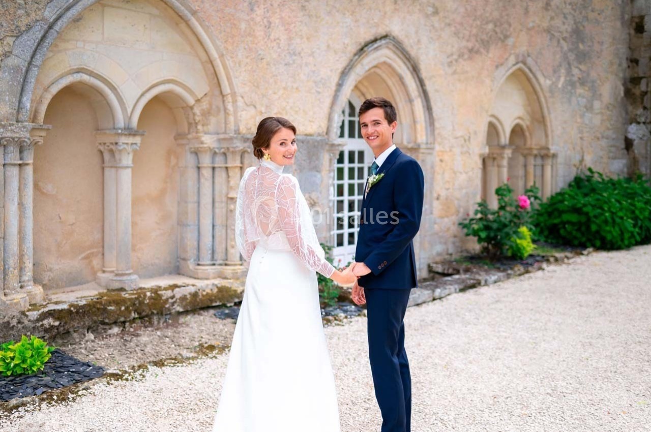 Un couple de mariés souriant se tient la main devant un mur en pierre avec des arches décoratives.