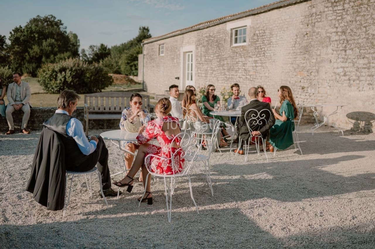 Personnes assises à des tables en plein air devant un bâtiment en pierre, discutant par une journée ensoleillée.