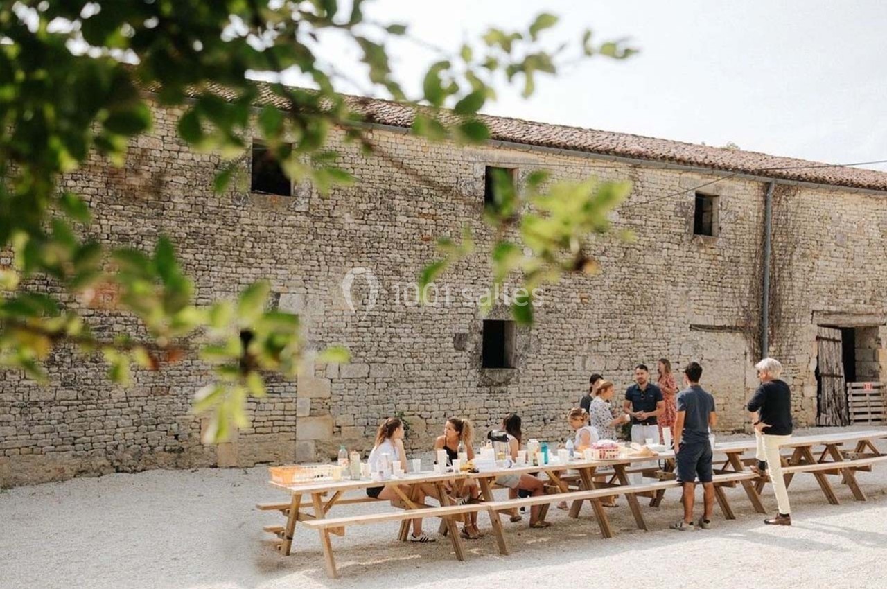 Groupe de personnes assises à des tables en bois, partageant un repas en extérieur devant un bâtiment en pierre.