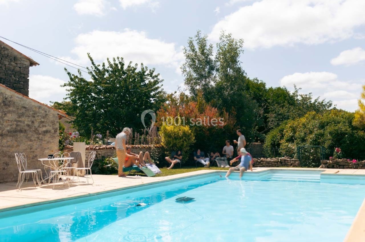 Personnes profitant d'une piscine extérieure dans un jardin ensoleillé avec des arbres et des buissons en arrière-plan.