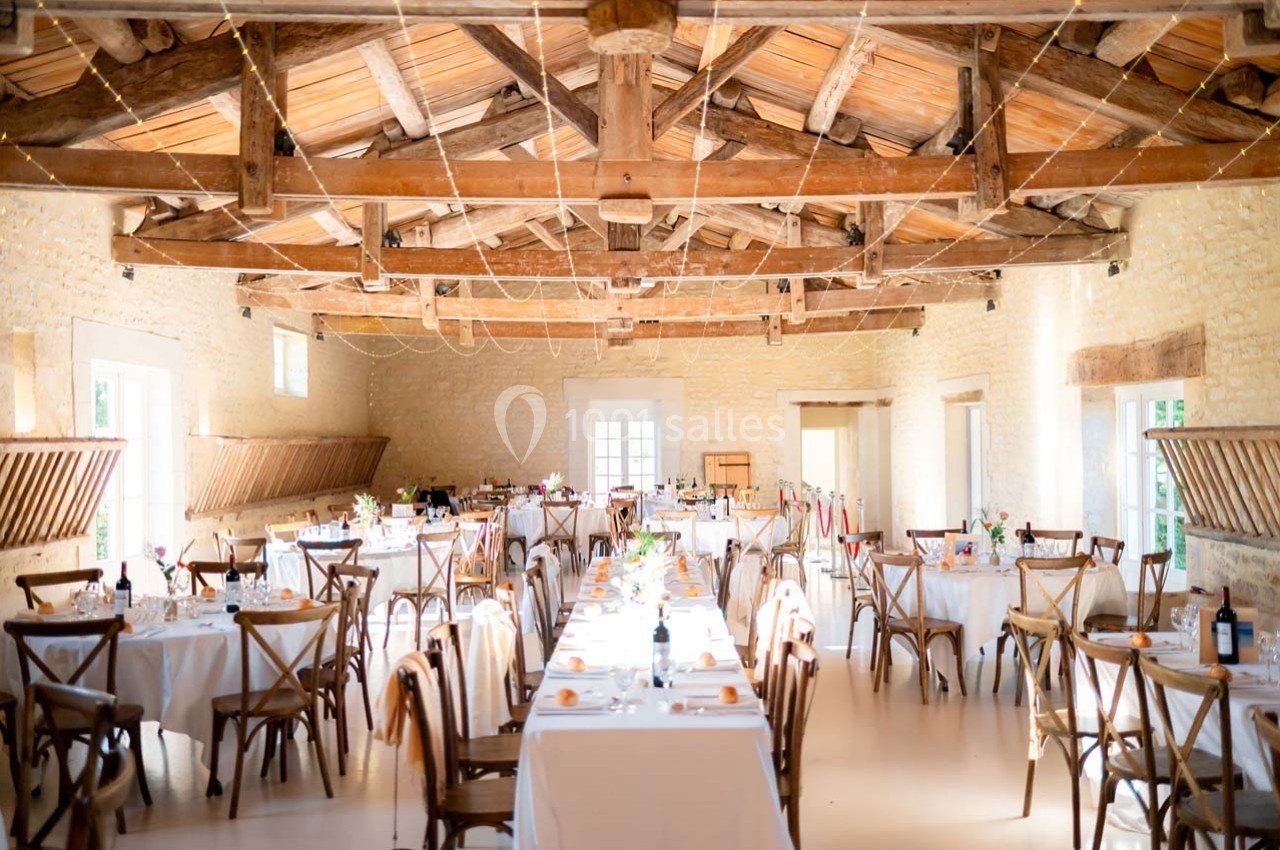 Salle de réception lumineuse avec tables dressées, chaises en bois et poutres apparentes au plafond.