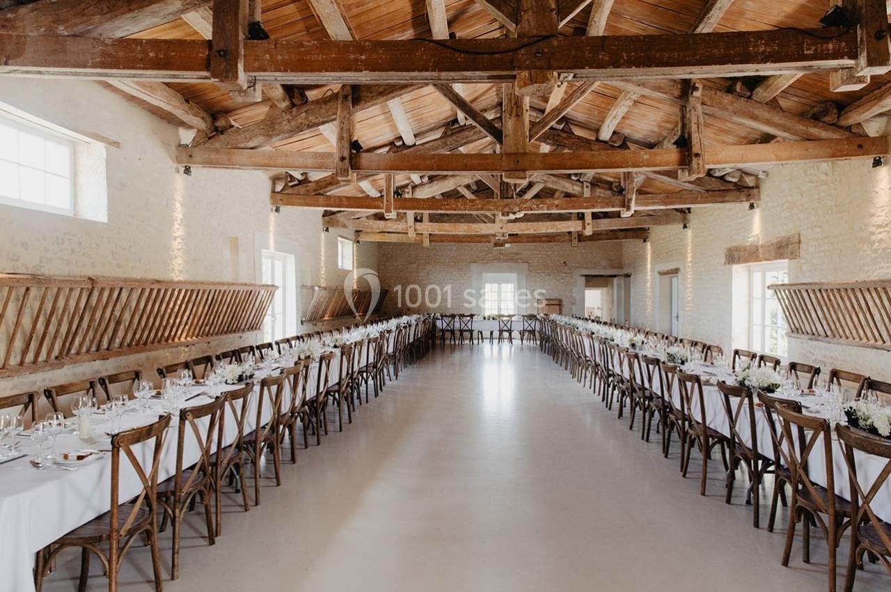 Salle de réception lumineuse avec poutres apparentes, tables longues dressées et chaises en bois alignées.