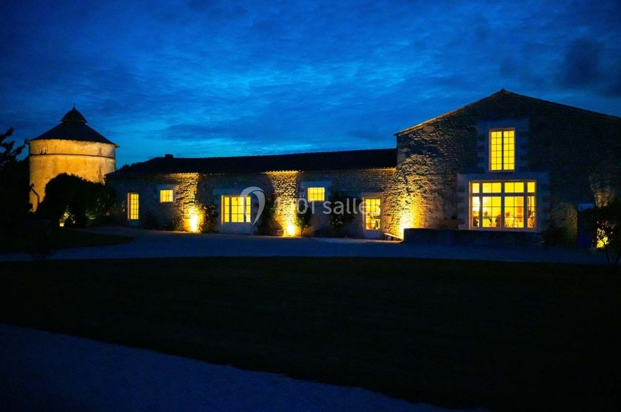 Bâtiment en pierre éclairé la nuit, avec un ciel bleu foncé en arrière-plan et un jardin sombre au premier plan.
