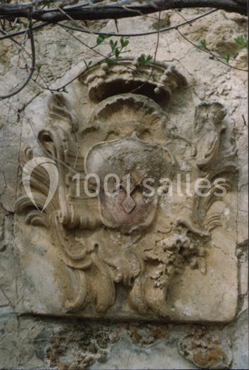 Blason sculpté en pierre, usé par le temps, fixé sur un mur ancien avec des branches visibles au-dessus.