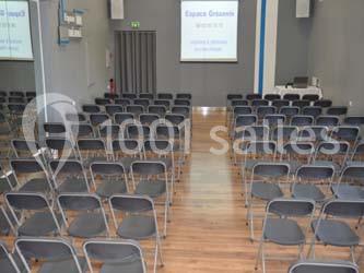 Salle de conférence avec rangées de chaises pliantes noires face à un écran de projection allumé.