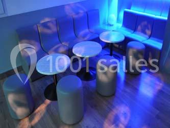 Banquettes grises, tables rondes et poufs dans une pièce éclairée par une lumière bleue tamisée.