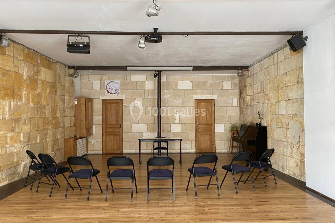Salle avec murs en pierre, sol en bois, chaises noires disposées en cercle et un bureau au fond.
