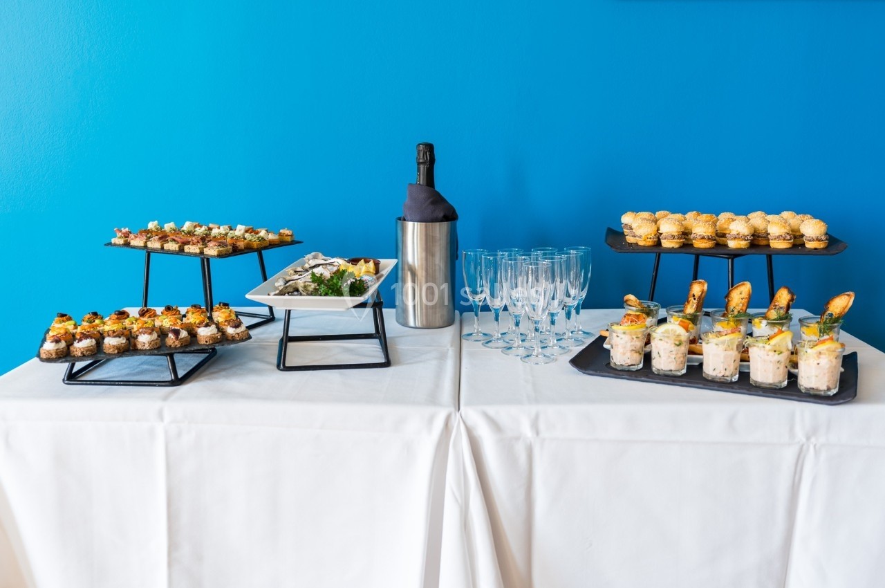 Buffet avec verrines, amuse-bouches variés, bouteille de champagne et flûtes sur une nappe blanche devant un mur bleu.