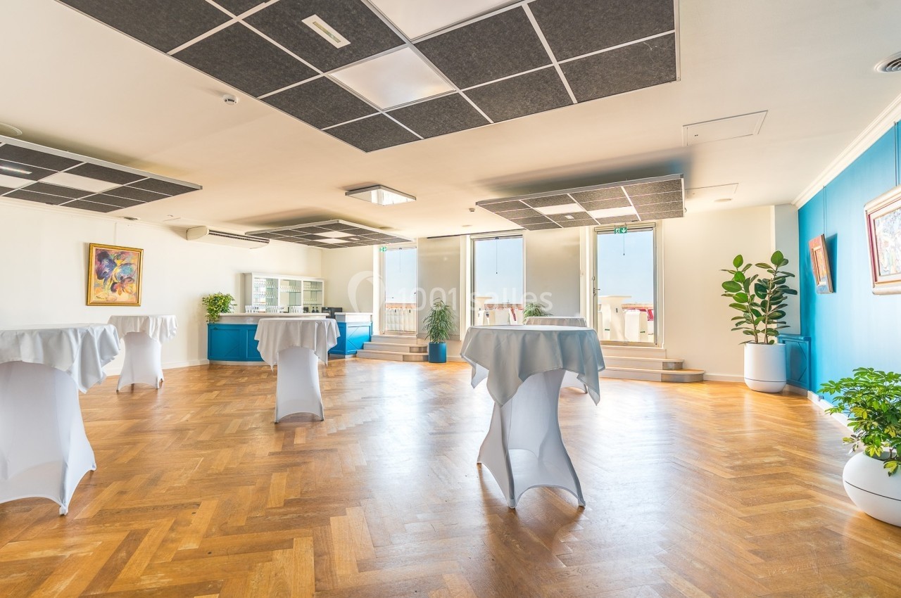 Salle lumineuse avec parquet en bois, tables hautes couvertes de nappes blanches, plantes et décorations murales.