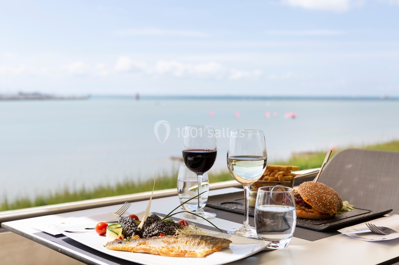 Repas en terrasse avec vue sur la mer, comprenant un plat de poisson, un burger, et des verres de vin et d'eau.