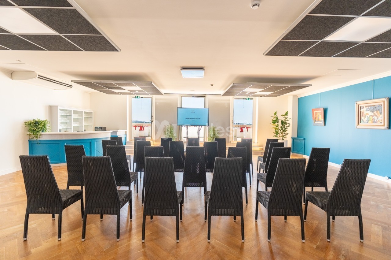 Salle de conférence lumineuse avec des chaises noires alignées face à un écran et un comptoir à l'arrière.