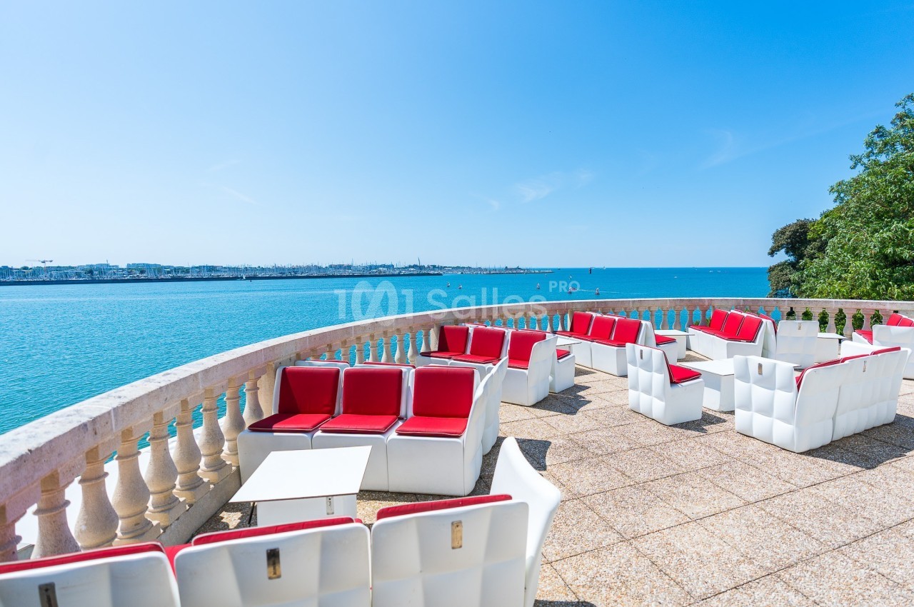 Terrasse ensoleillée avec fauteuils rouges et blancs, vue sur une mer calme et un horizon dégagé.