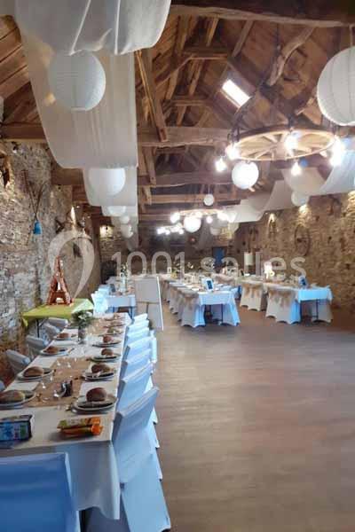 Salle de réception rustique avec tables dressées, nappes blanches et décorations suspendues sous un plafond en bois.