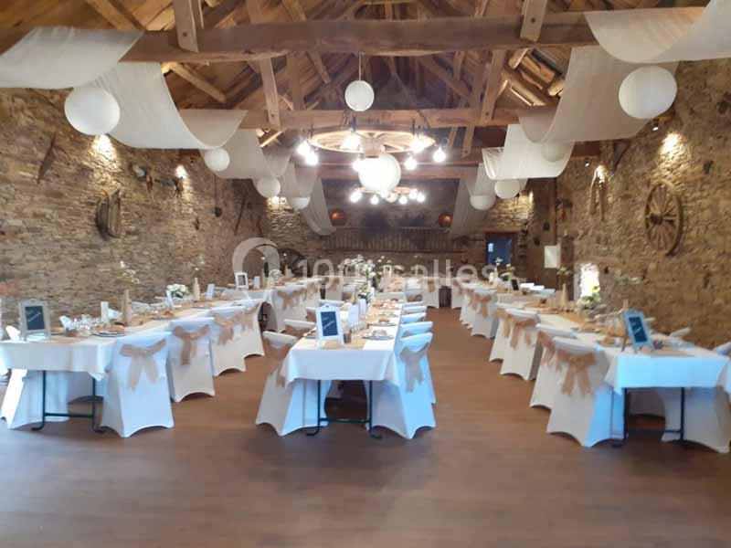 Salle de réception rustique décorée pour un événement, avec tables dressées et nappes blanches sous un plafond en bois.