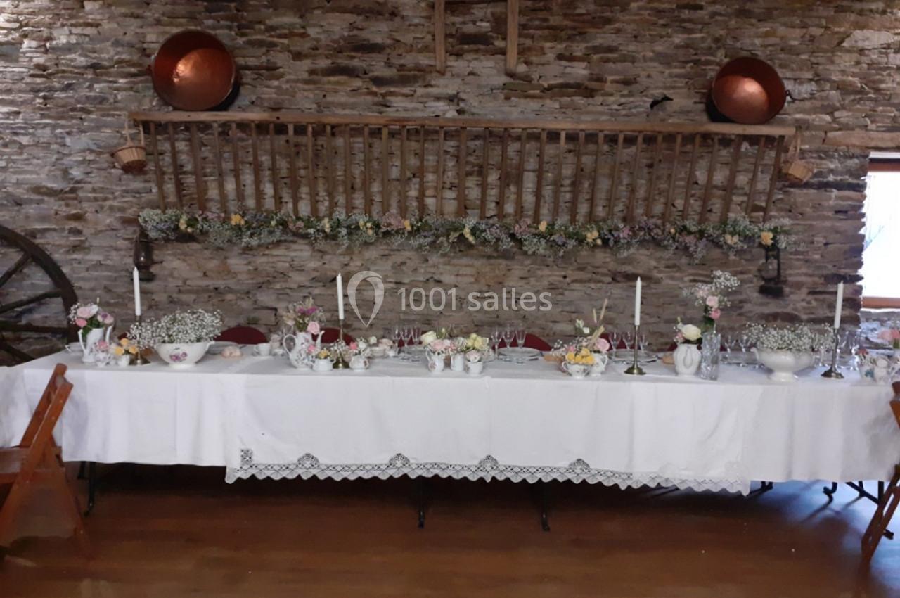 Table décorée pour un événement, avec nappes blanches, fleurs, bougies et vaisselle, dans une salle en pierre rustique.