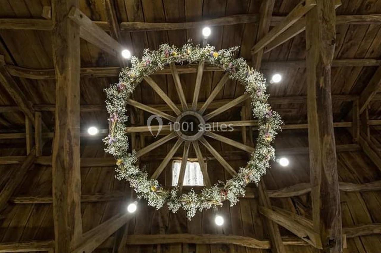 Vue d'une charpente en bois avec une roue décorée de fleurs suspendue au plafond, éclairée par des spots lumineux.