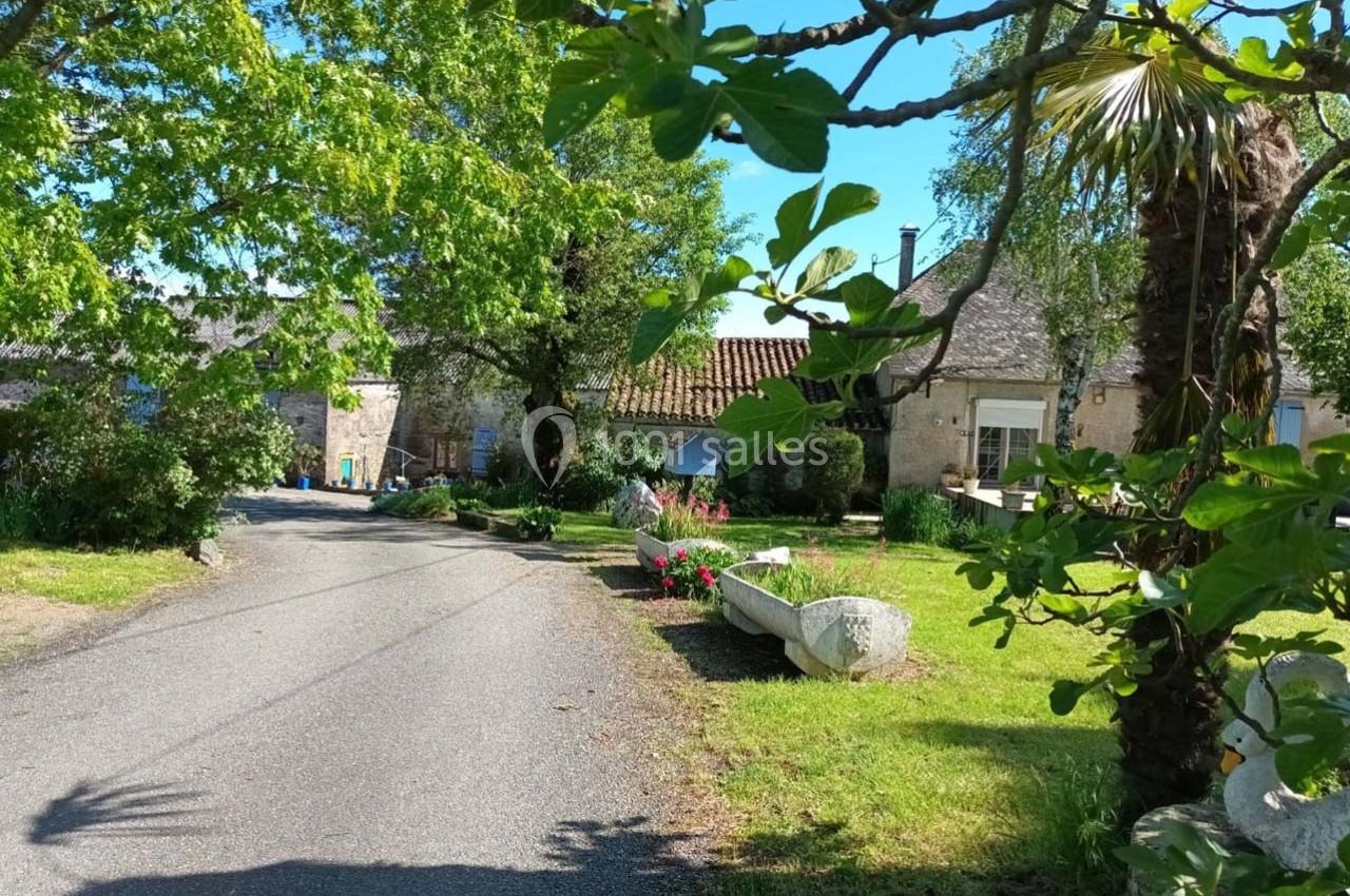 Chemin bordé de verdure menant à des maisons en pierre sous un ciel bleu, avec arbres et fleurs en premier plan.