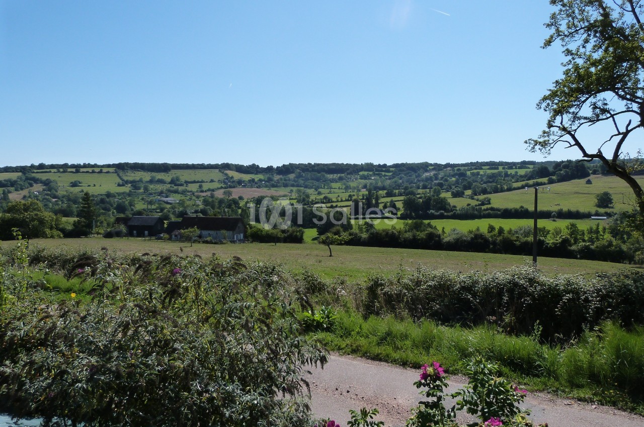 Paysage rural avec champs verdoyants, haies, quelques maisons et arbres sous un ciel bleu dégagé.