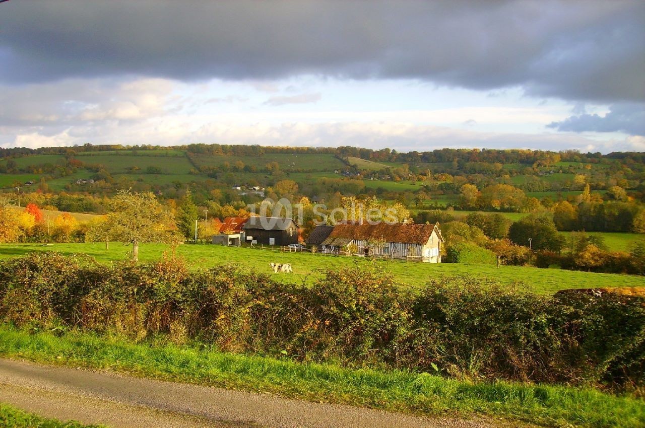 Paysage rural avec une ferme traditionnelle entourée de champs verdoyants et d'arbres sous un ciel nuageux.