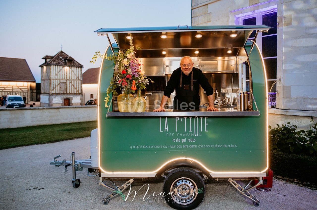 Un homme souriant se tient dans un food truck vert décoré de fleurs, stationné devant un bâtiment éclairé.