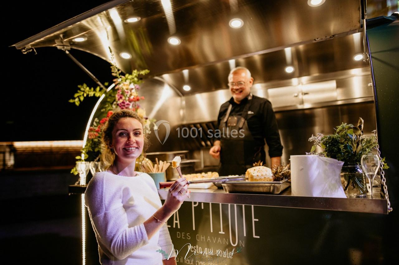 Une femme souriante tenant un verre devant un food truck éclairé, avec un cuisinier en arrière-plan.