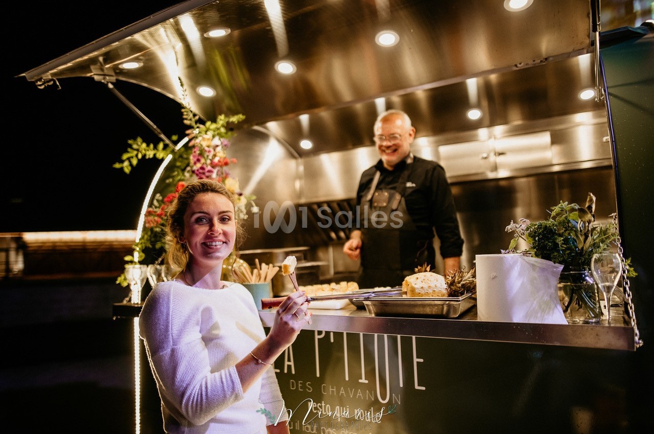 Une femme souriante tenant un verre devant un food truck éclairé, avec un cuisinier en arrière-plan.
