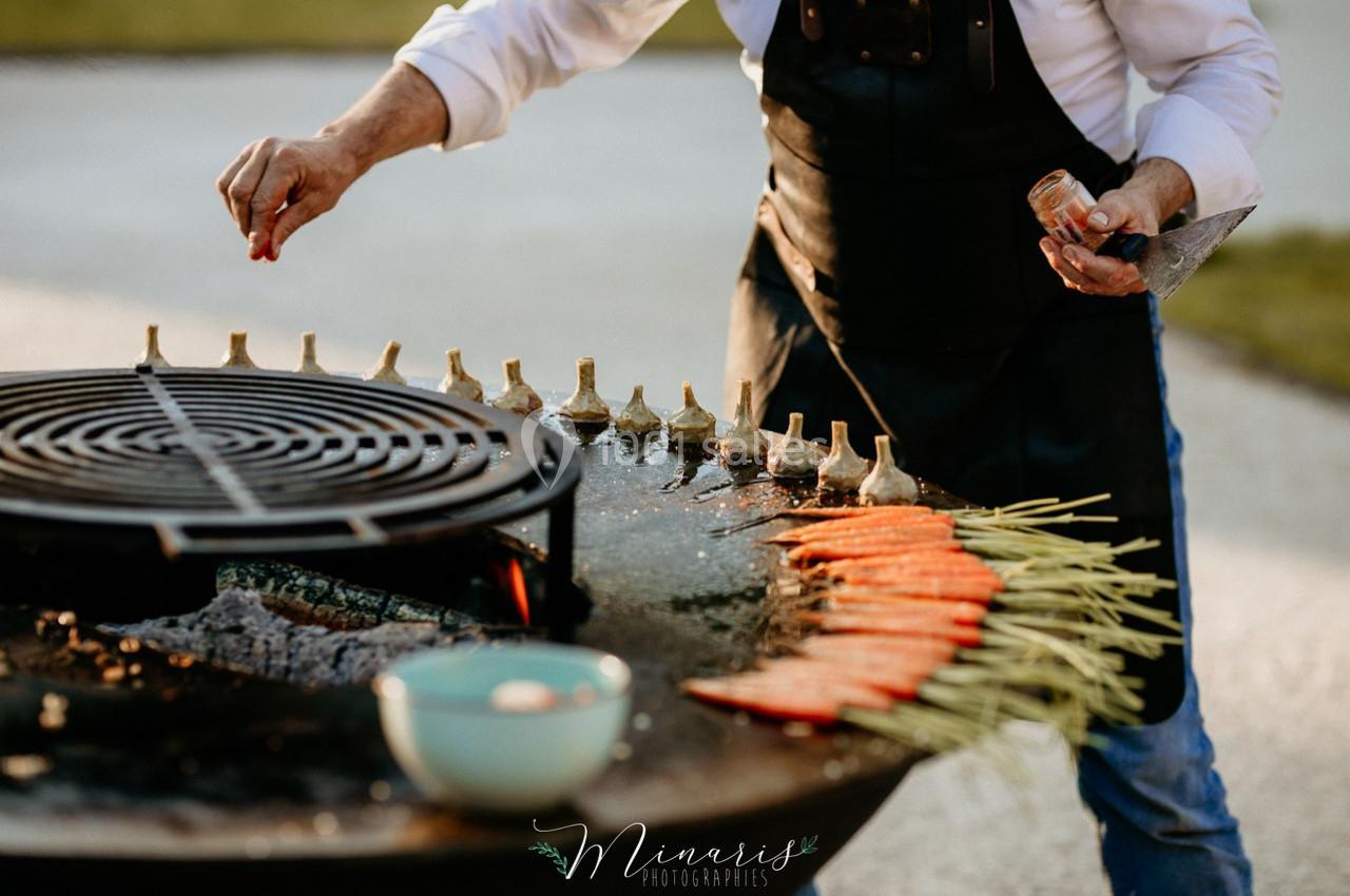 Un cuisinier assaisonne des légumes grillés, dont des carottes et des artichauts, sur une plancha en plein air.