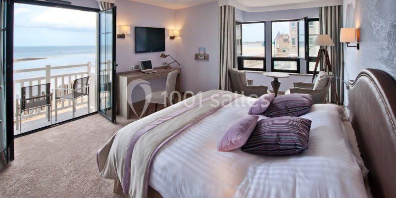 Chambre lumineuse avec lit double, bureau, balcon ouvert et vue sur la mer et la plage.