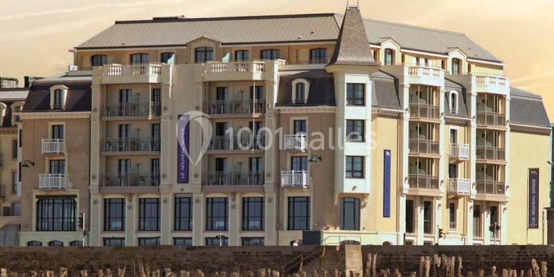Façade d'un grand bâtiment beige avec balcons et toits mansardés, situé près d'une plage.