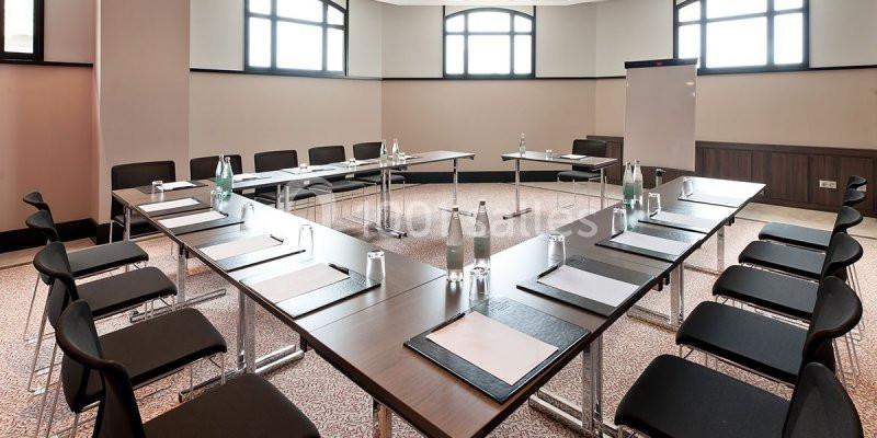 Salle de réunion lumineuse avec tables disposées en U, chaises noires, bouteilles d'eau et tableau blanc.