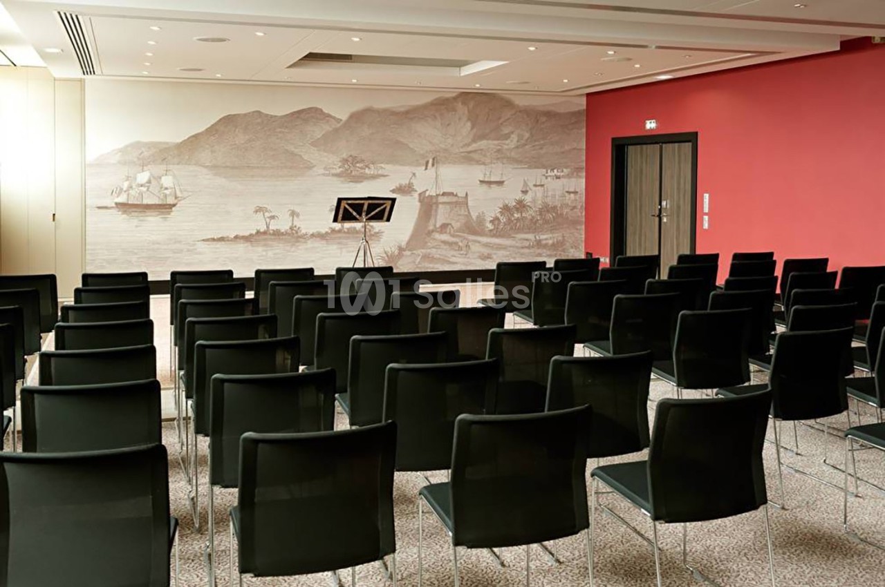 Salle de conférence vide avec des rangées de chaises noires, un mur rouge et une fresque représentant un paysage maritime.